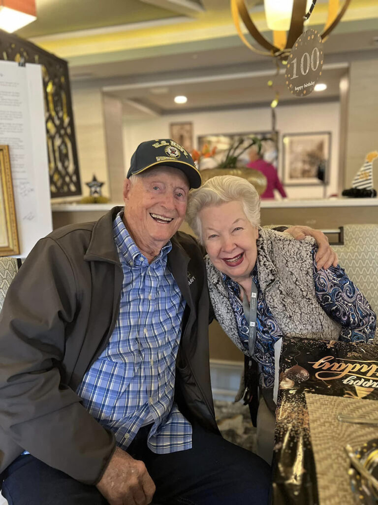 Smiling older couple posing in front of a table, celebrating a joyful 100th birthday milestone.