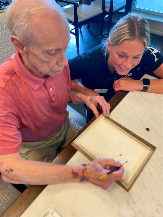 Elderly man painting with a brush on a framed canvas, while a smiling young woman watches at The Plaza at Wildwood Senior Living.