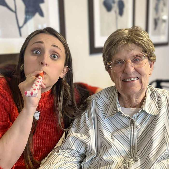 Senior resident smiles while seated next to a playful team member in a red sweater blowing a party horn, creating a fun and cheerful atmosphere during a community celebration.