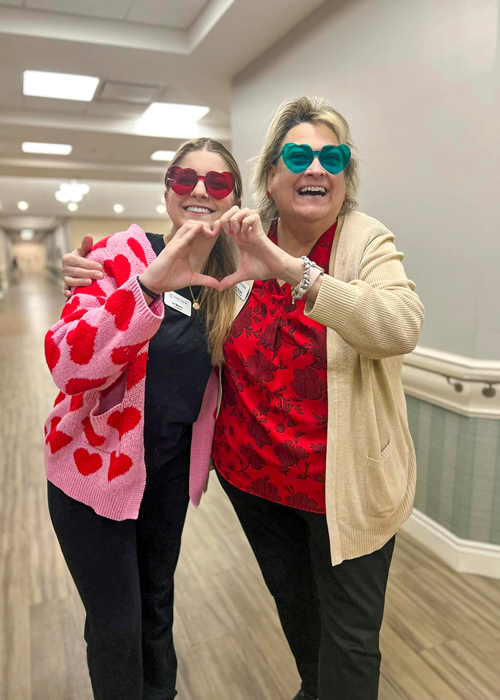 Two senior living staff members, dressed in festive heart-themed outfits and heart-shaped sunglasses, smiling and making a heart shape with their hands to celebrate Valentine's Day.