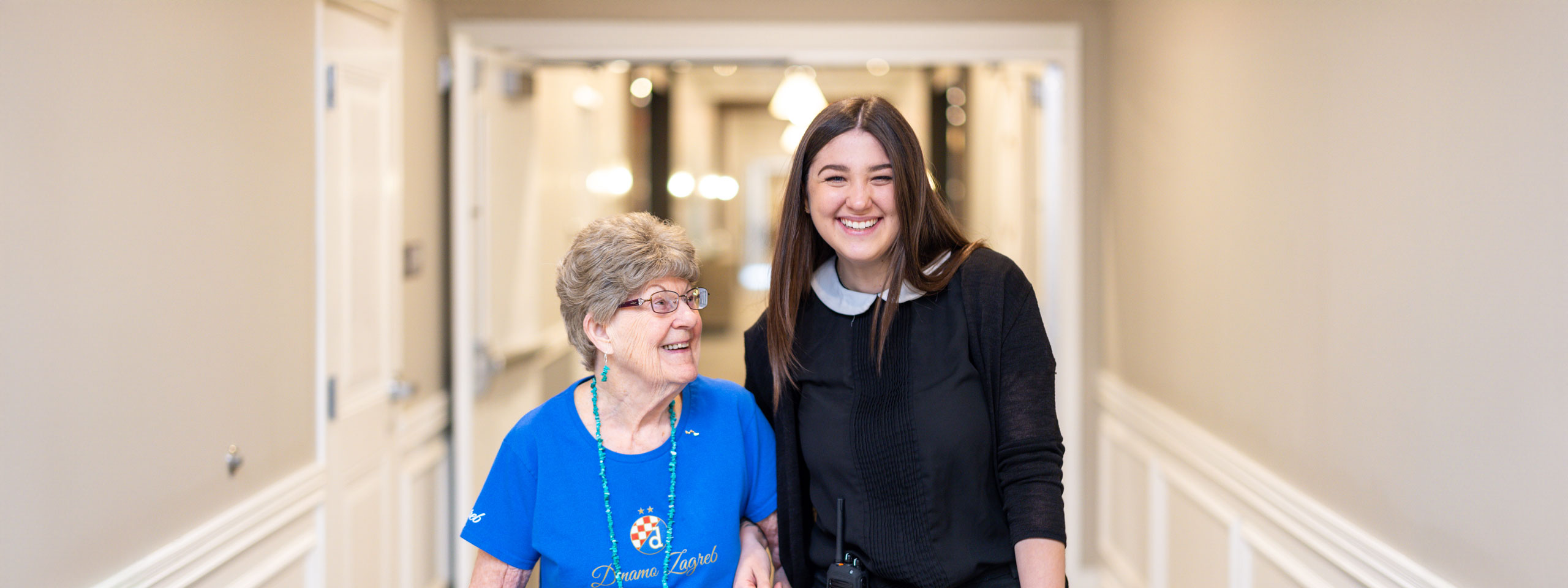 Senior resident wearing a blue shirt smiles while walking down a hallway with a young, friendly staff member, showcasing a warm and caring atmosphere in a senior living community.