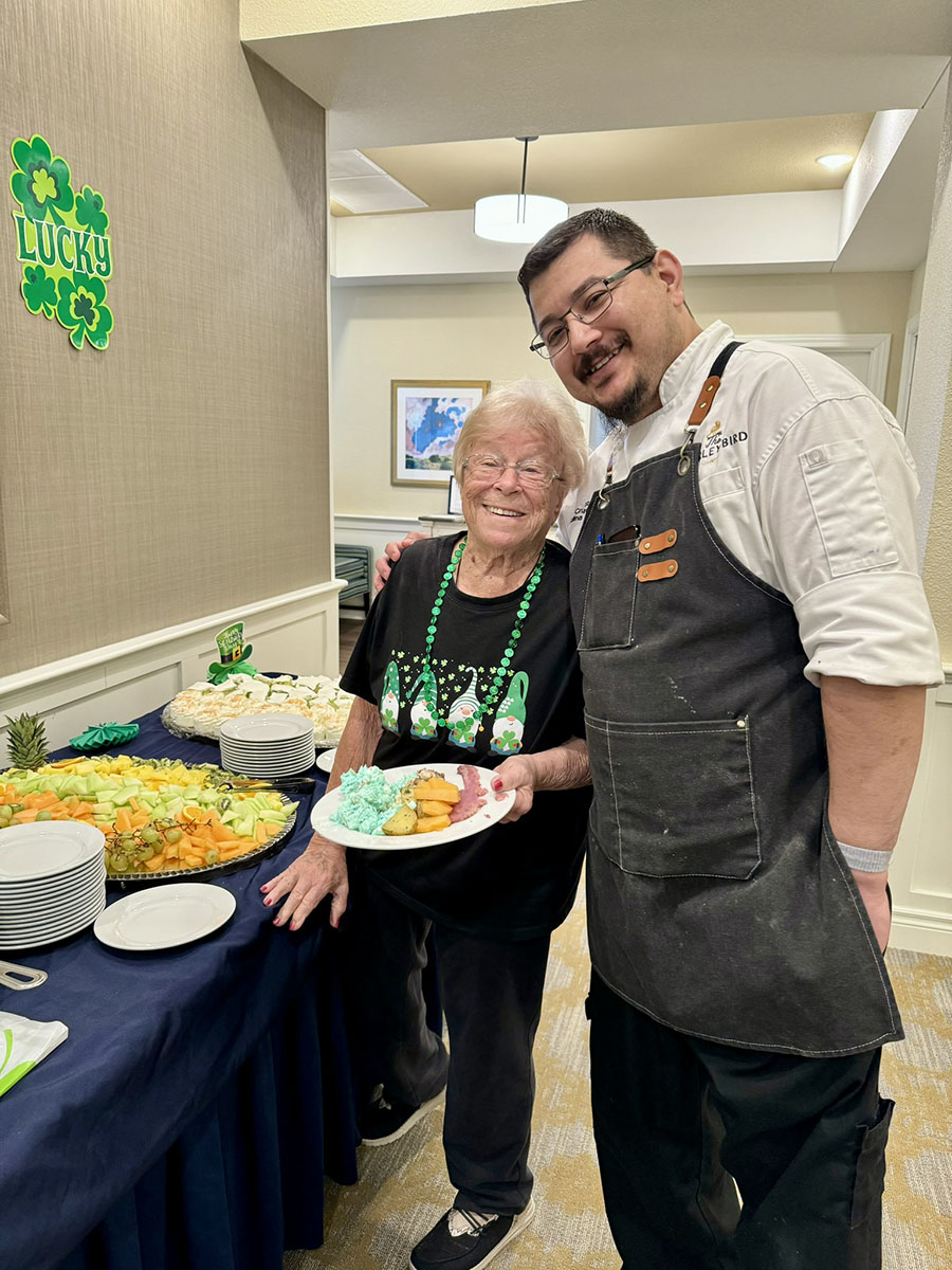 Senior resident in festive St. Patrick's Day attire holding a plate of food, smiling next to a chef in a white uniform, standing by a buffet table decorated with fruit and desserts.