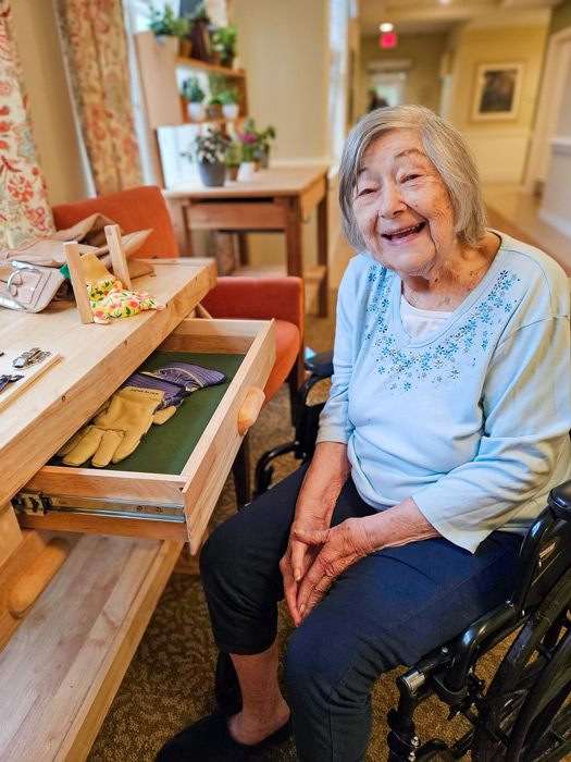 A senior resident smiling while sitting in a wheelchair beside a wooden workbench with open drawers, gardening tools, and gloves, enjoying an activity in a senior living community.