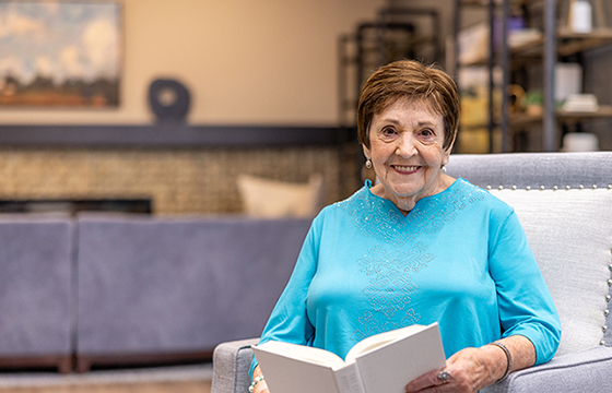 A senior resident wearing a turquoise blouse sits comfortably in a cozy, modern living room, smiling warmly while reading a book, with soft furnishings and artwork in the background.