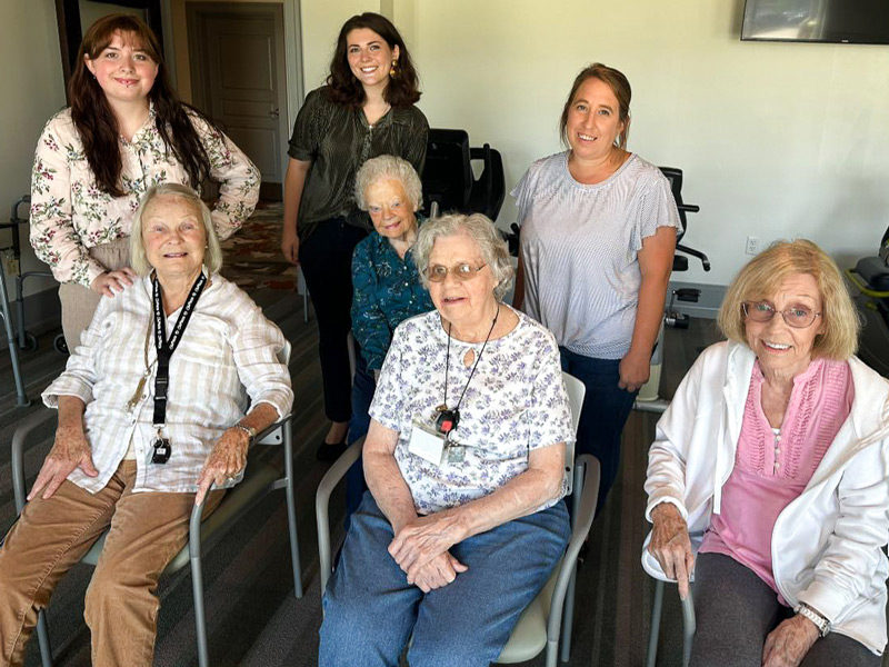 A group of smiling senior residents and staff members gather together, seated and standing, sharing a joyful moment during a community event.