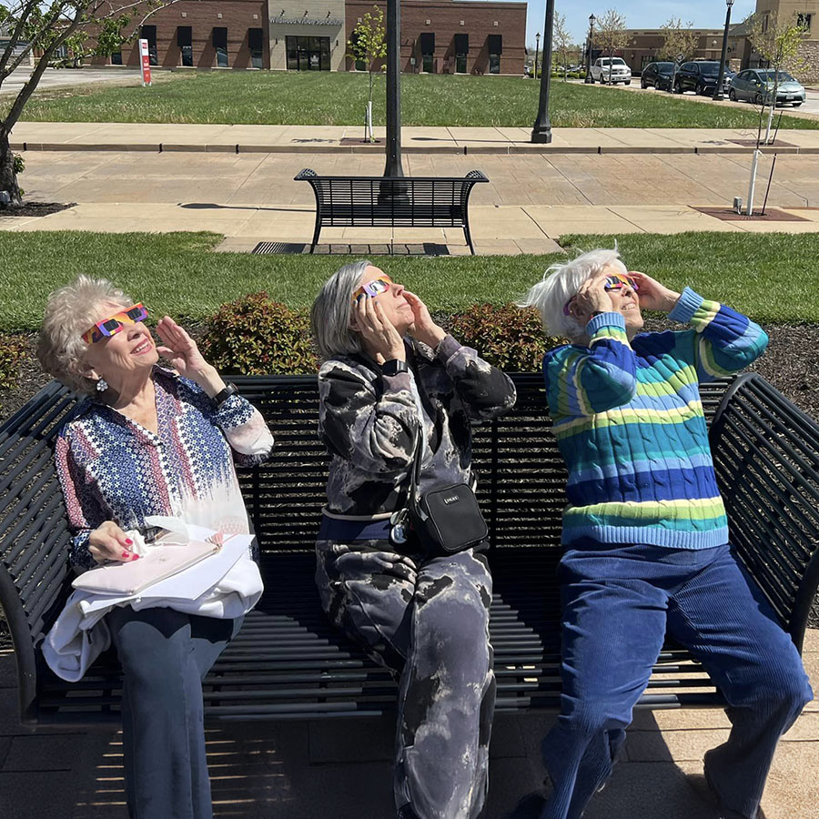 Three senior resident women enjoying the moment, watching the eclipse with safety sunglasses while seated on a bench, at the 2024 Eclipse Watching Party in The Plaza at Wildwood Senior Living.