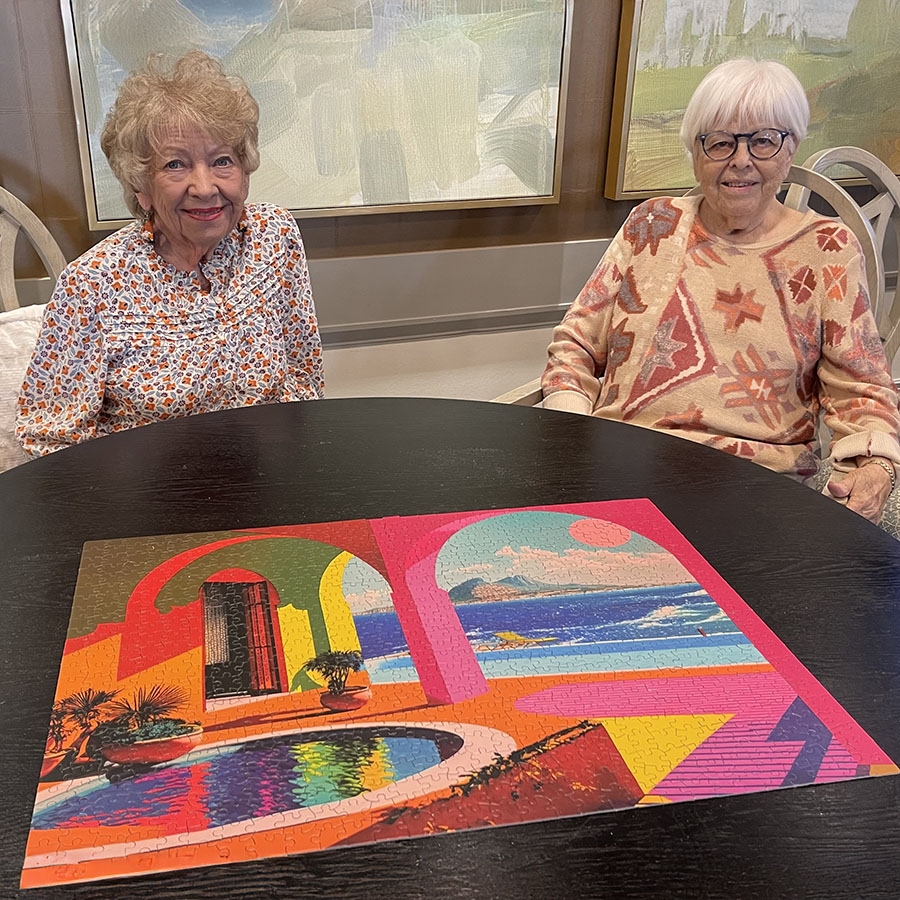 Two elderly women smiling and sitting at a table, celebrating their accomplishment of completing a puzzle together.