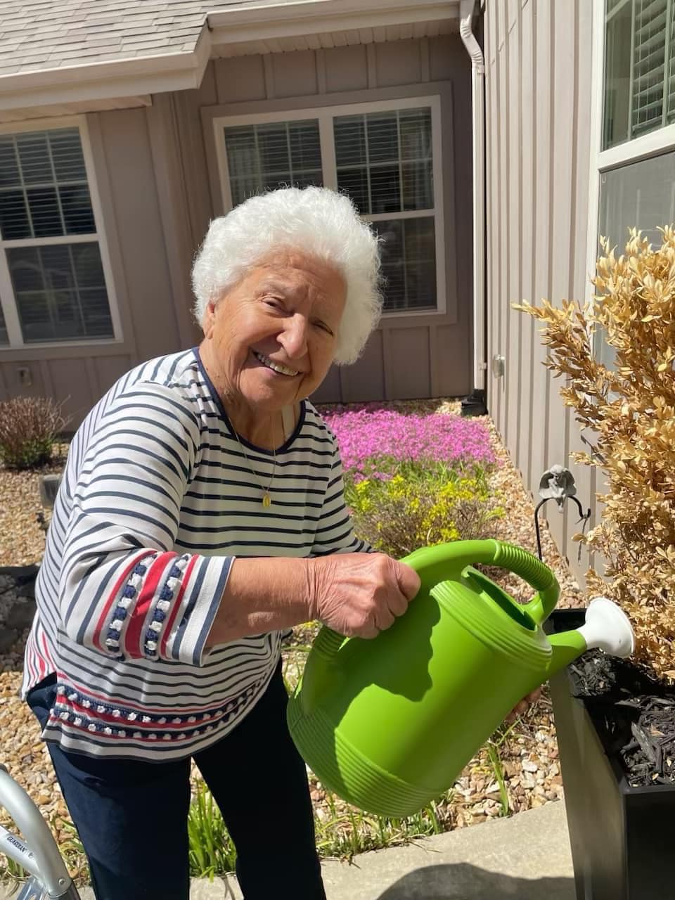 A senior resident with white hair smiles while watering plants outdoors with a green watering can, enjoying a sunny day in the garden at a senior living community.