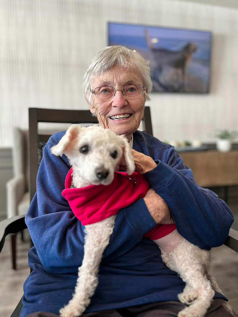 Senior resident smiling while holding a small white dog dressed in a red sweater. They are seated indoors with a framed picture of a dog in the background.