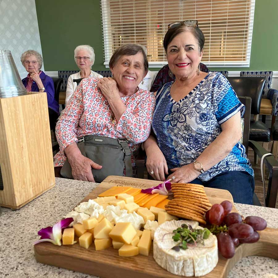 Two senior residents smile and pose together at a table with a beautifully arranged cheese and cracker platter, surrounded by friends enjoying a social gathering in a cozy setting.