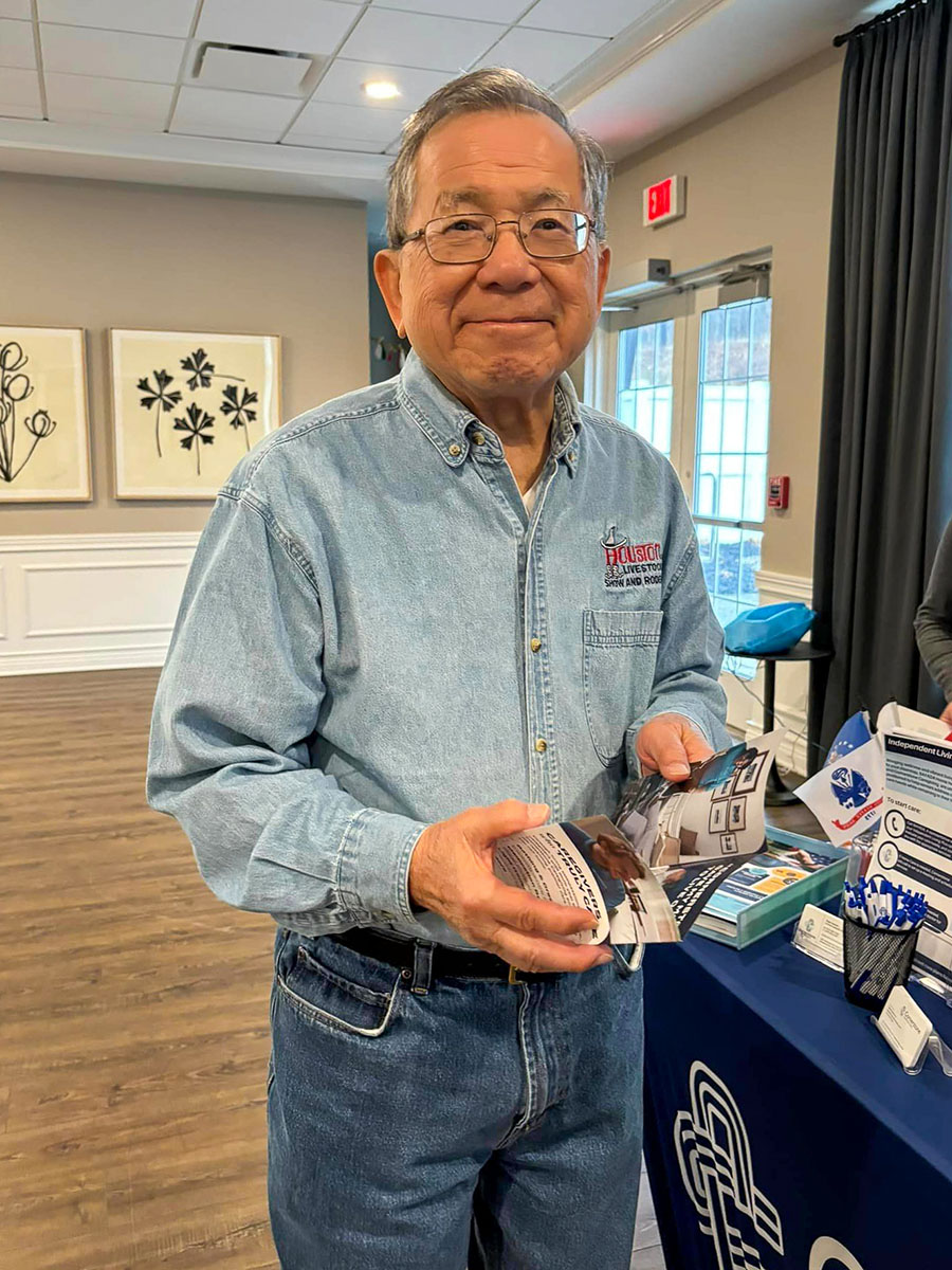 Smiling senior resident in a denim shirt holds informational brochures during a Wellness Wednesday event, standing in a well-lit room with tables displaying health and wellness materials.