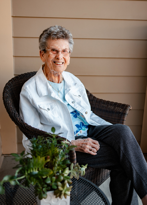 A smiling senior resident sitting comfortably on a wicker chair on the porch, dressed in a white jacket and floral shirt, enjoying a peaceful moment with a nearby potted plant.