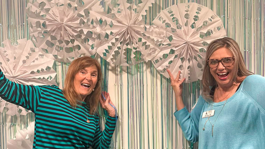 Two team members in front of a festive backdrop with large white snowflake decorations, smiling and posing playfully during a community winter event.