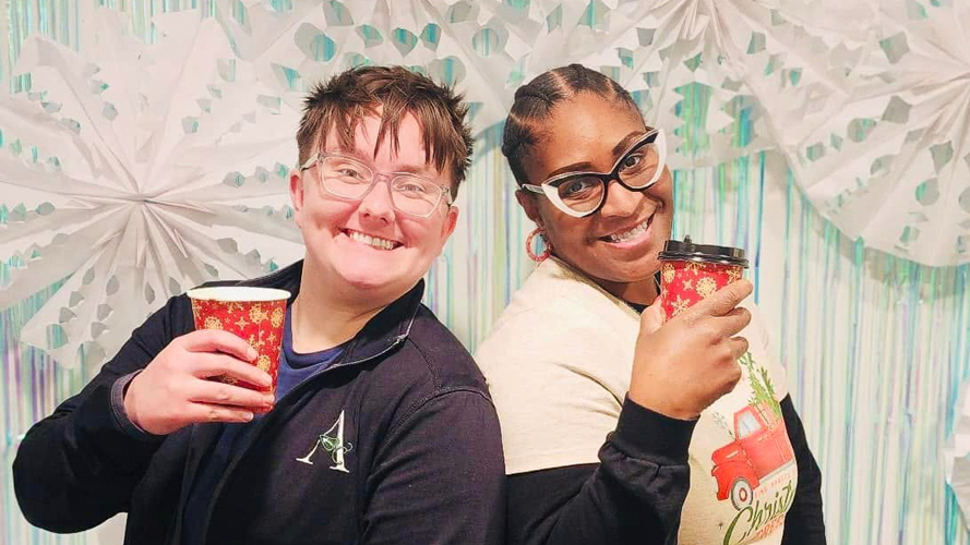 Two staff members holding holiday-themed coffee cups in front of a snowy-themed backdrop with large paper snowflakes during a festive gathering.