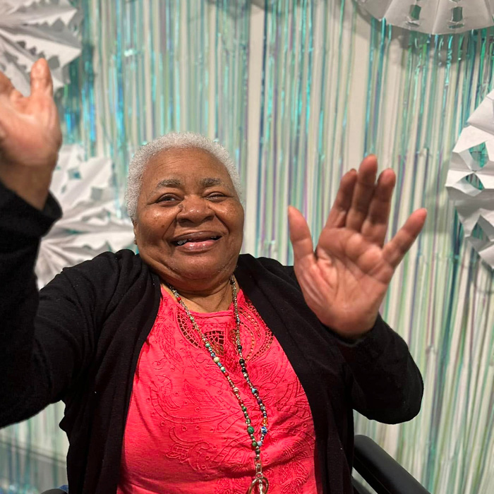 A senior resident in a bright pink blouse, raising her hands with a big smile, celebrating a holiday event in front of a snowy backdrop.