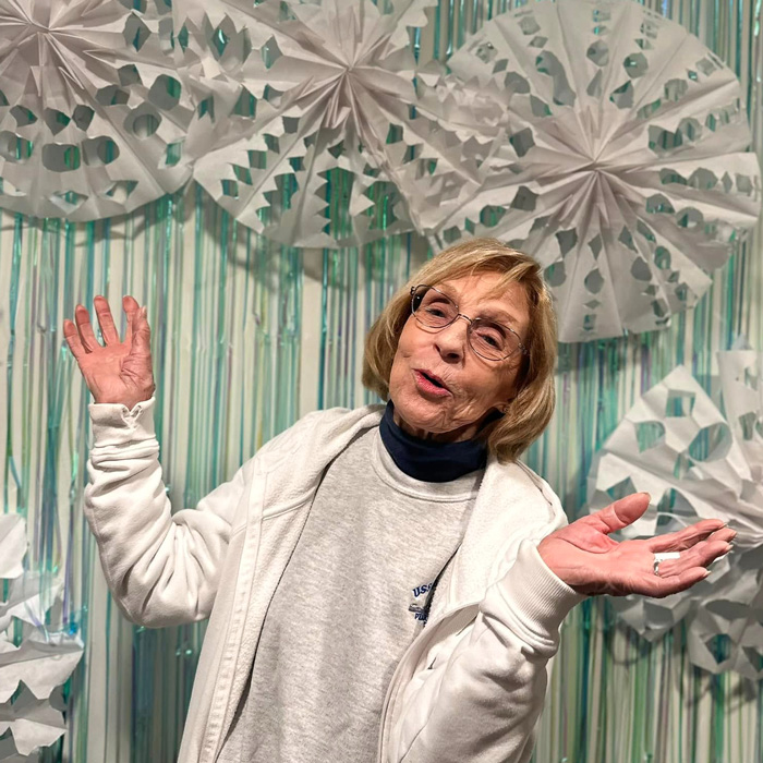 A senior resident gesturing joyfully with her hands in front of a festive backdrop featuring intricate paper snowflakes during a community winter event.